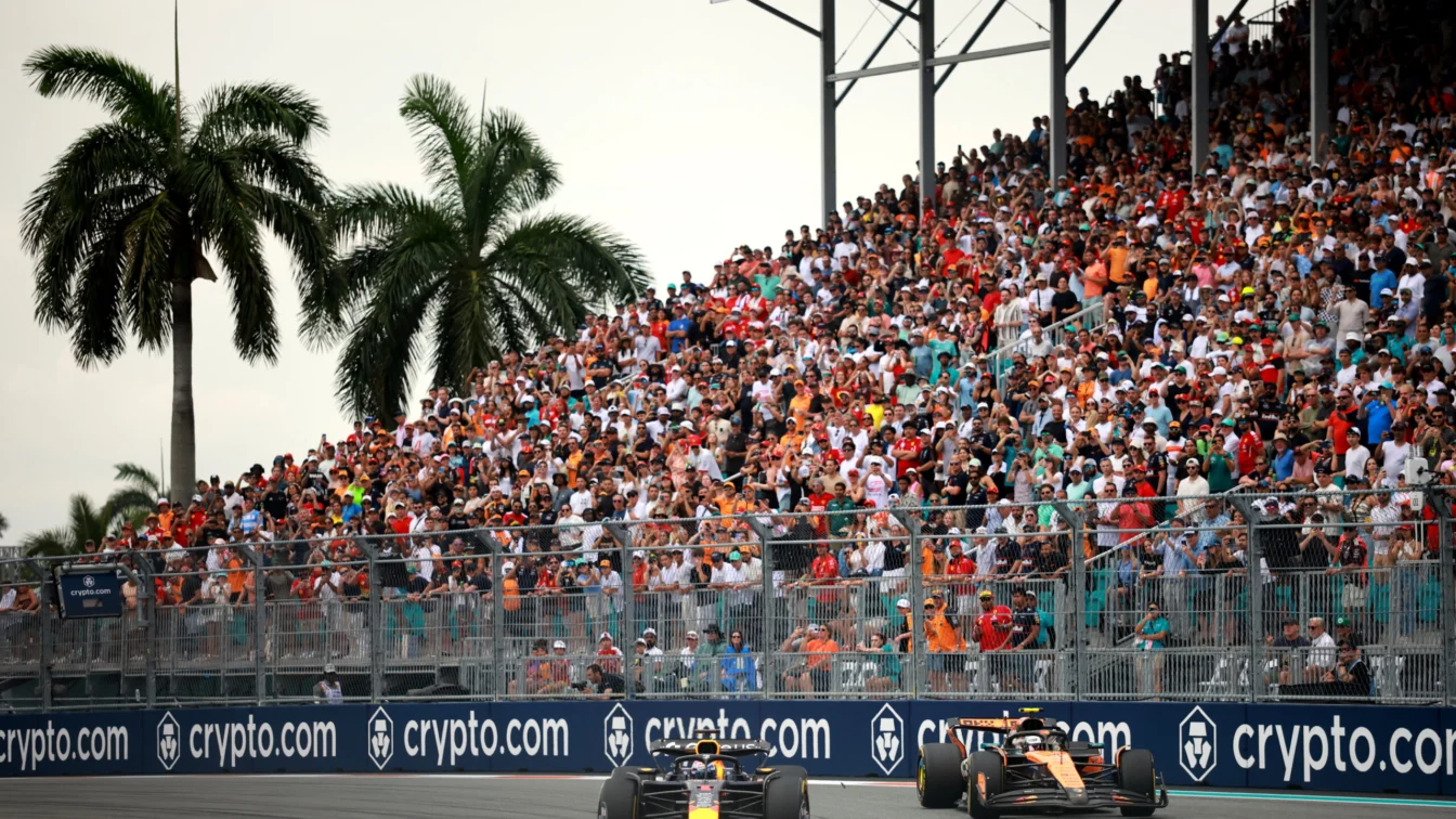 Max Verstappen leading Lando Norris in a Formula 1 race at Miami International Autodrome, with a crowd of spectators in the background.