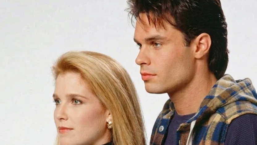 Two individuals, one male and one female, are standing side by side and posing for a photograph. The male individual is dressed in a jacket with a checkered pattern, while the female individual is wearing a high-neck top. They are both smiling and appear to be in a studio setting.