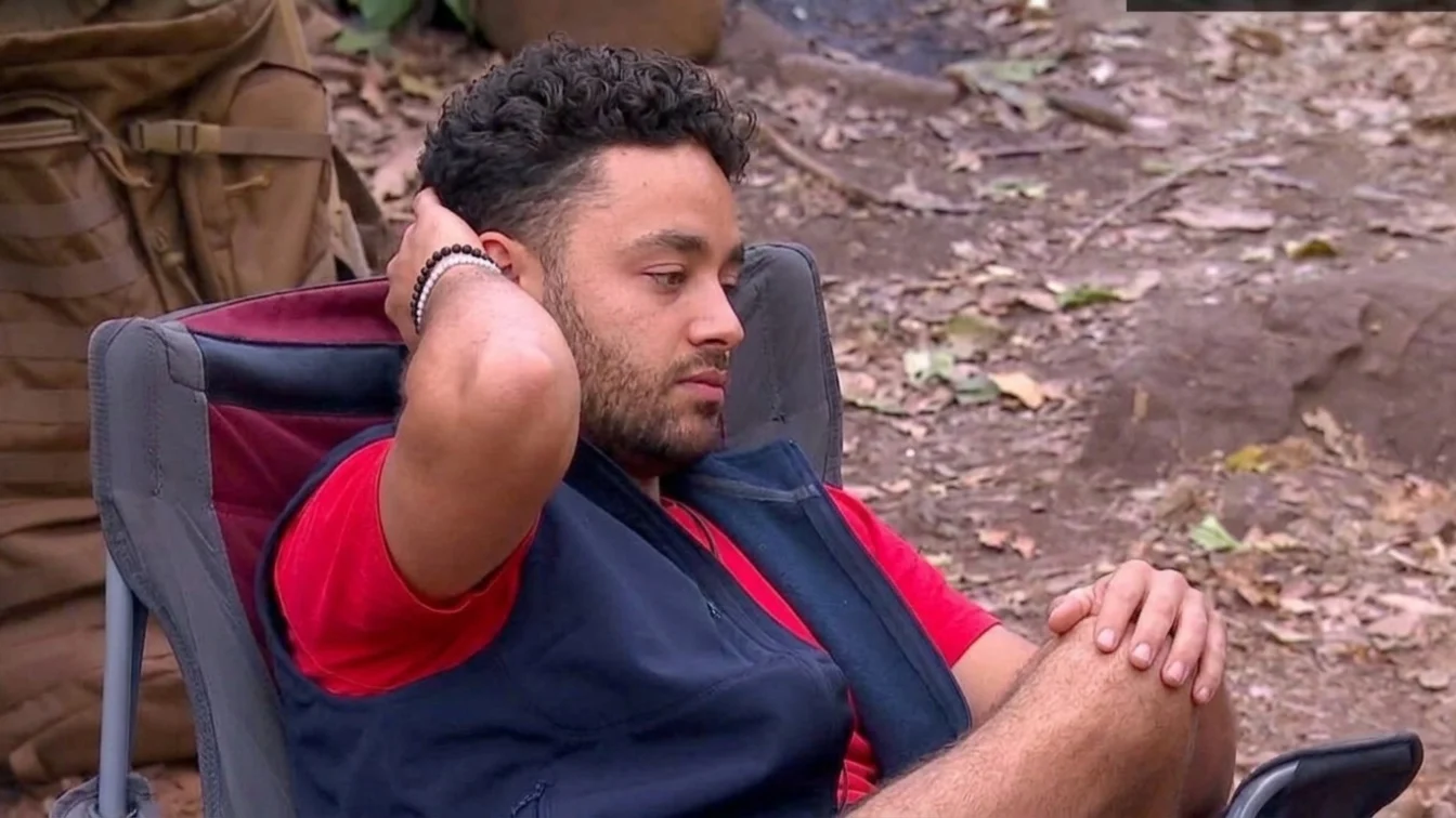 An individual dressed in a red and blue shirt, along with a vest, is reclining on the ground. Their head is resting on their left hand, while their right hand is positioned on their knee. The setting appears to be an outdoor area with rocks and dirt.