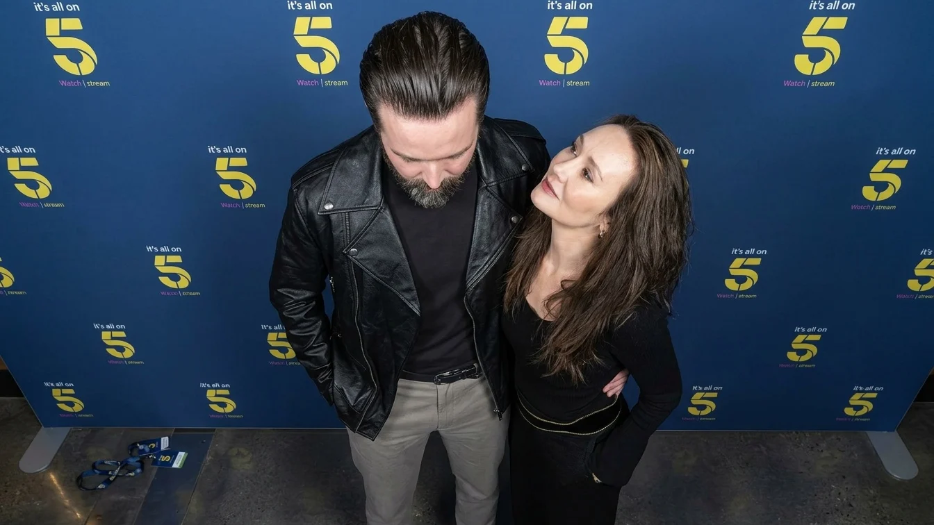 An adult male and an adult female are standing side by side in front of a blue backdrop with yellow text and numbers.