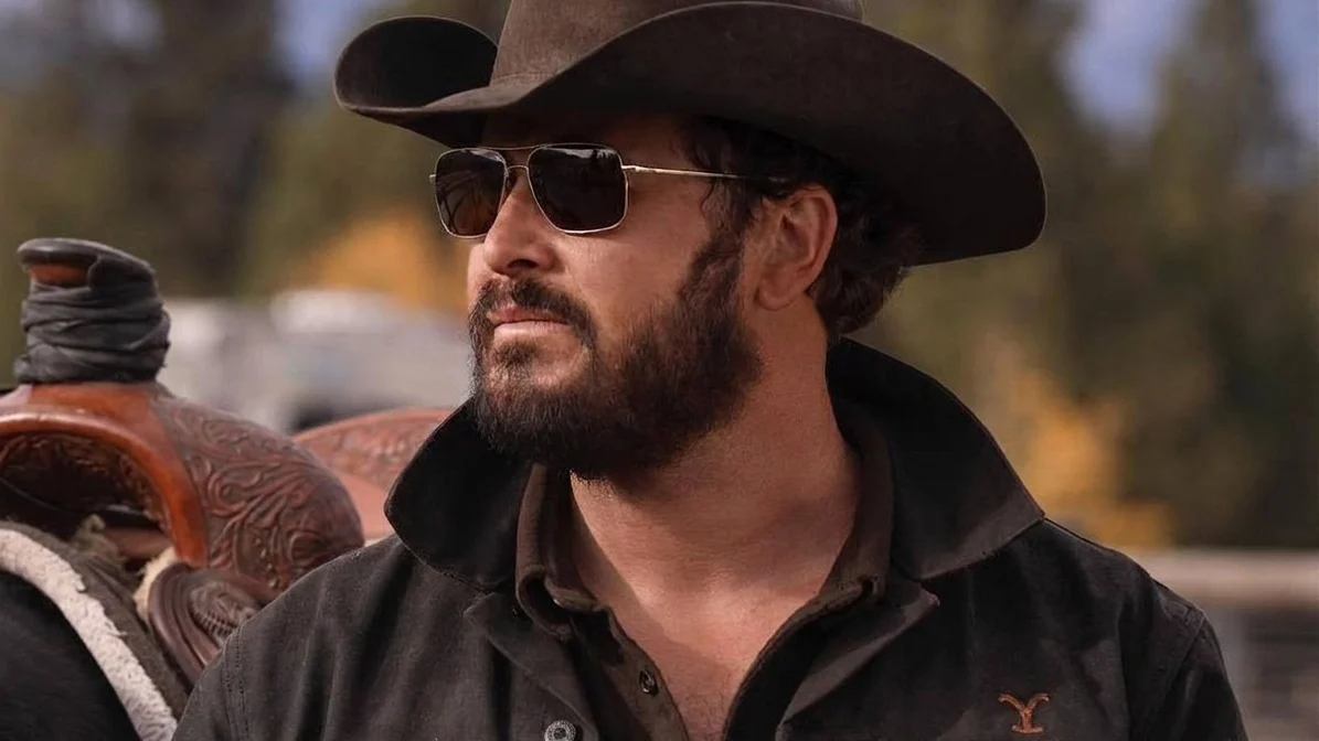 An individual with a beard, donning a black jacket and a cowboy hat, is standing in an outdoor setting with a blurred background featuring trees and other individuals.
