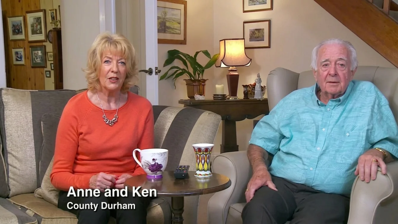 Two individuals, a man and a woman, are seated on a couch within a domestic setting. The woman is dressed in a red long-sleeved top, while the man is clad in a blue long-sleeved shirt. They are positioned in close proximity to a table that holds various items, including a cup and a small potted plant. The backdrop features additional furniture and decorative elements, such as a lamp and picture frames.