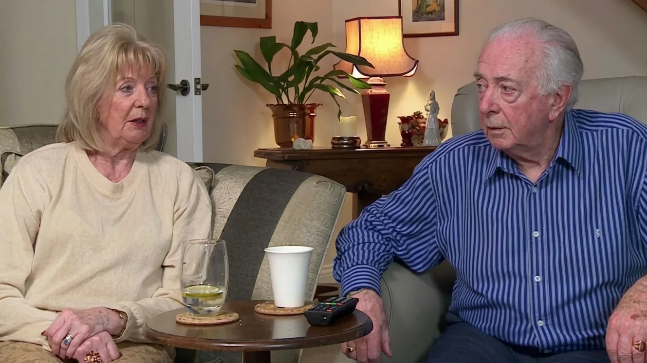 An elderly couple is sitting together on a couch in a cozy living room, with a small table in front of them holding a cup and a remote control. The room is adorned with various decorative items, including a potted plant and a lamp on a side table.