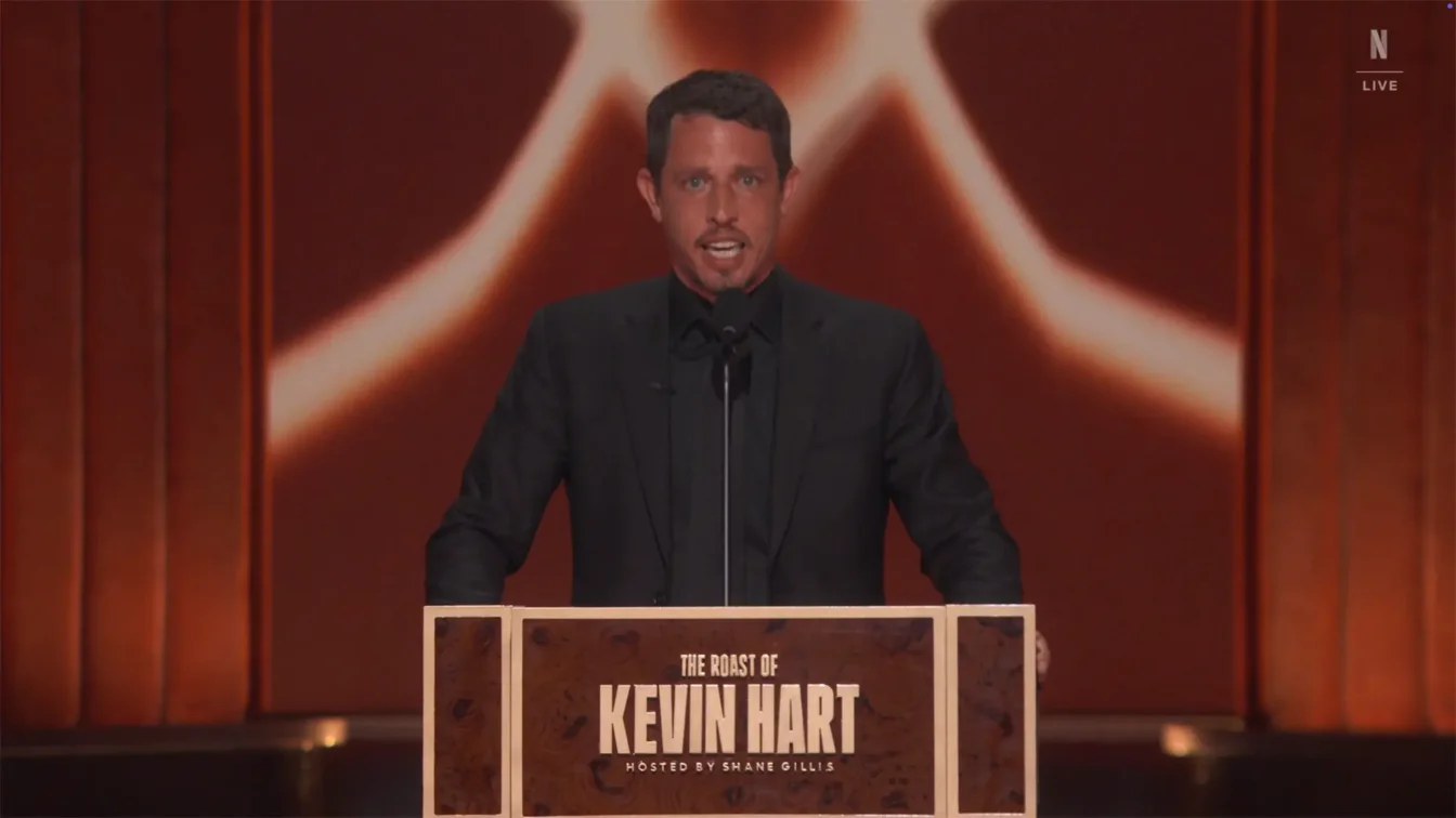 Tony Hinchcliffe standing at a podium labeled "The Roast of Kevin Hart" on a red stage with an abstract background.