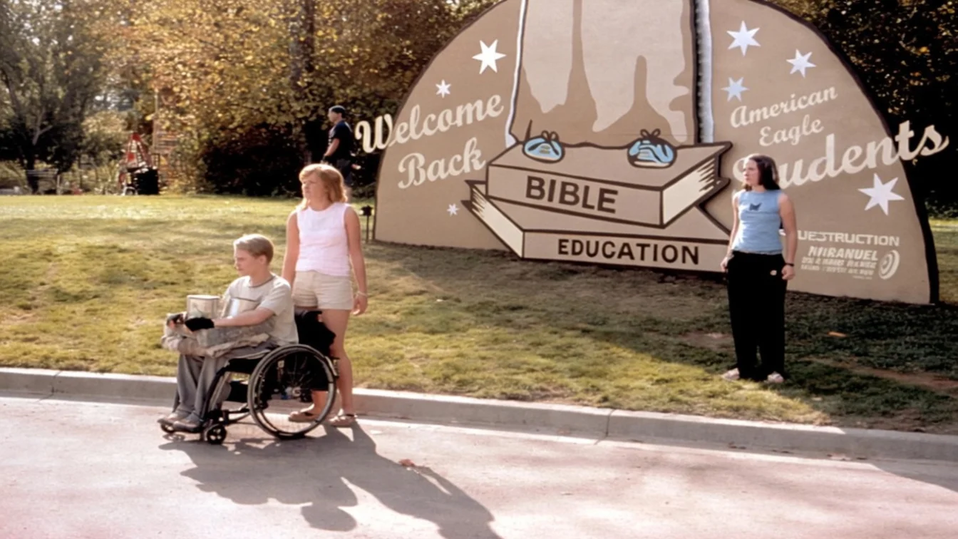 Macaulay Culkin in a wheelchair, Mandy Moore, and Jena Malone stand next to a "Welcome Back American Eagle Students, Bible Education" sign in the movie Saved.