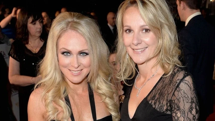 Two women with blonde hair, dressed in black evening gowns, smiling and posing for a photograph at an event, with blurred figures in the background.