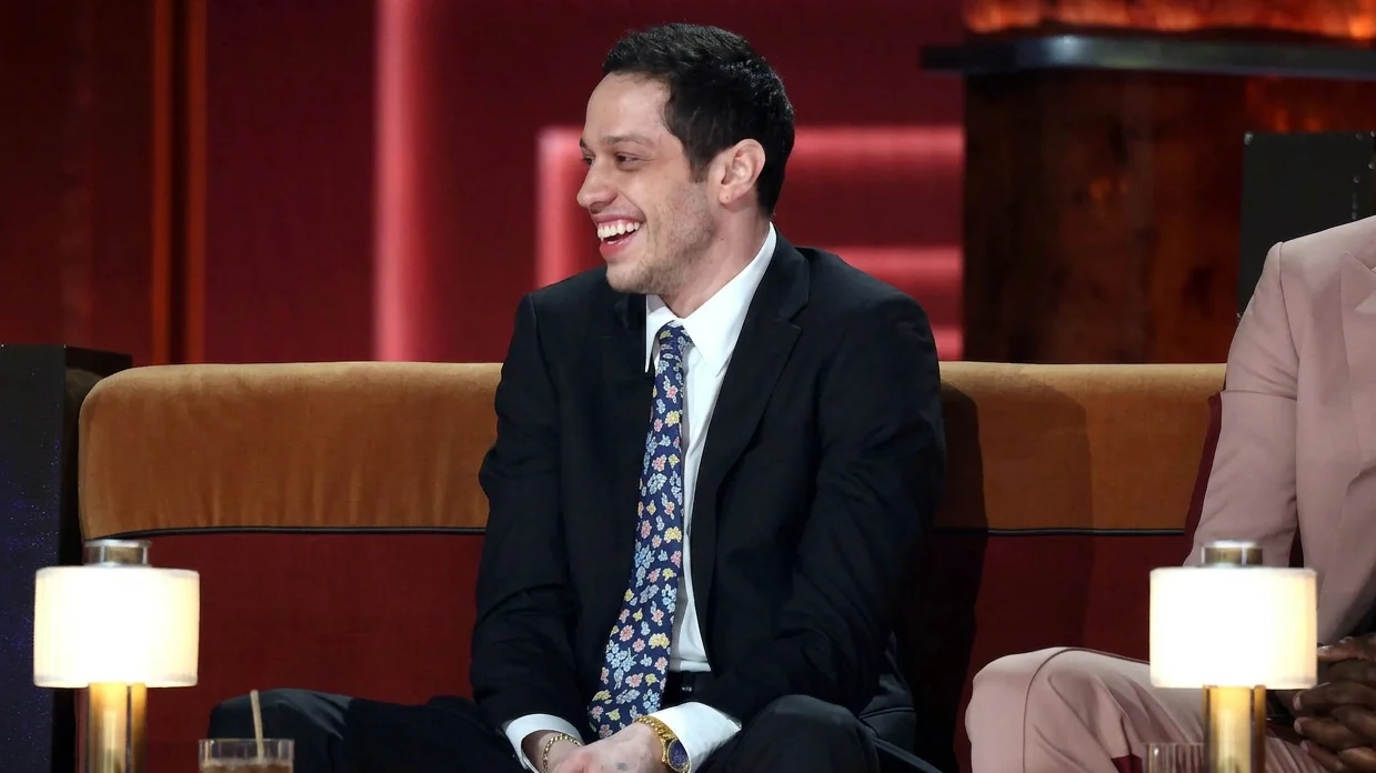 Pete Davidson smiling while wearing a black suit and a floral tie.