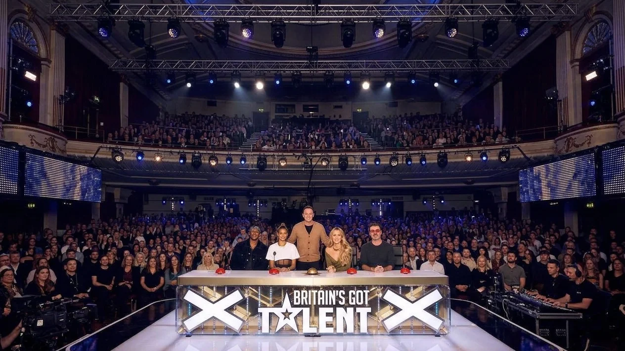A group of individuals, both male and female, are standing behind a table in a dimly lit room. They are likely posing for a photograph with a crowd visible in the background.