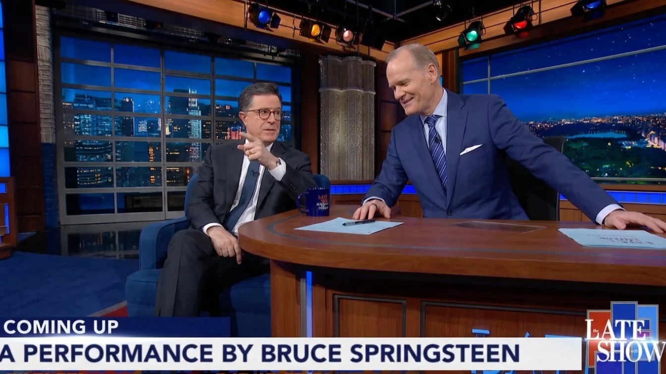 Stephen Colbert pointing at the viewer next to Jeff Glor at The Late Show desk.