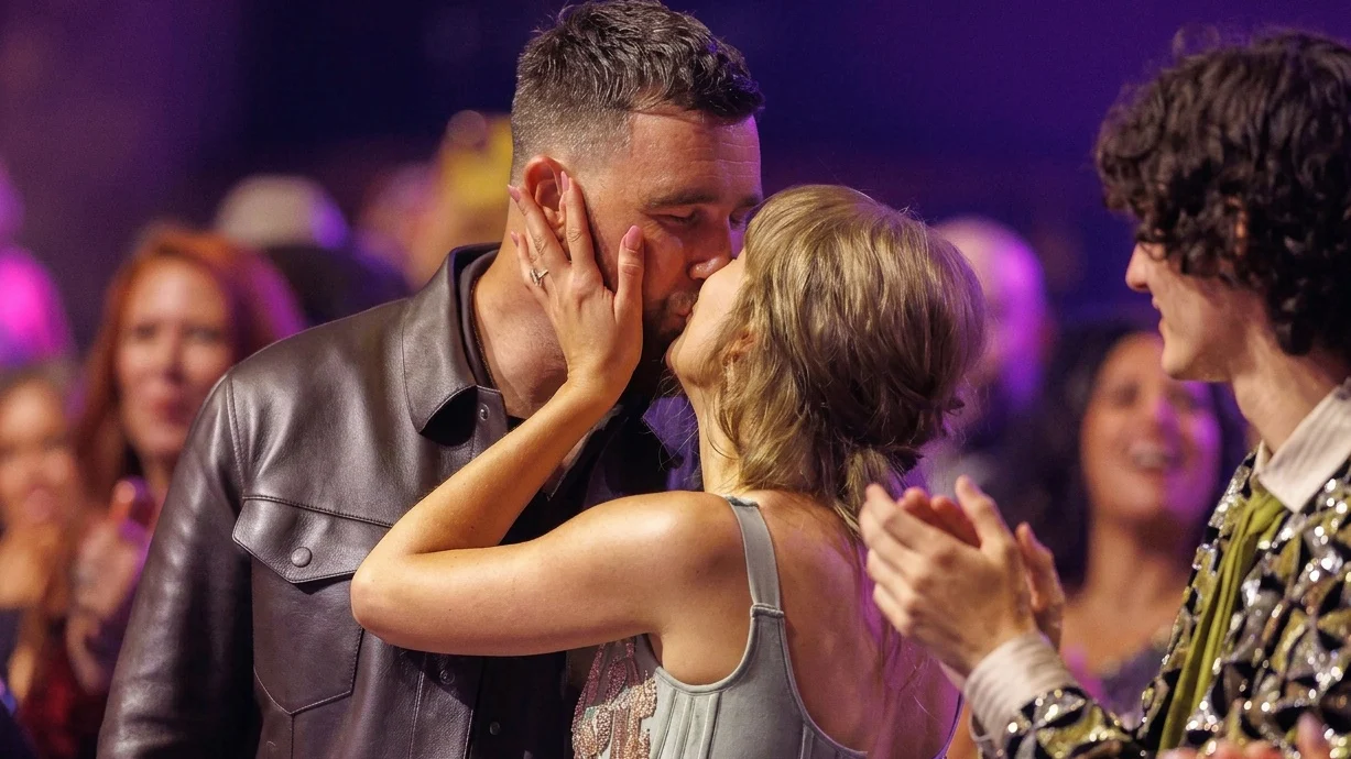 Taylor Swift, in a grey mini dress, kissing Travis Kelce, in a brown leather jacket.