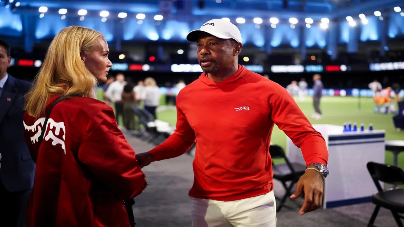 Tiger Woods, wearing a red long-sleeved shirt and white cap, speaks to his girlfriend, Vanessa Trump, who wears a red jacket.