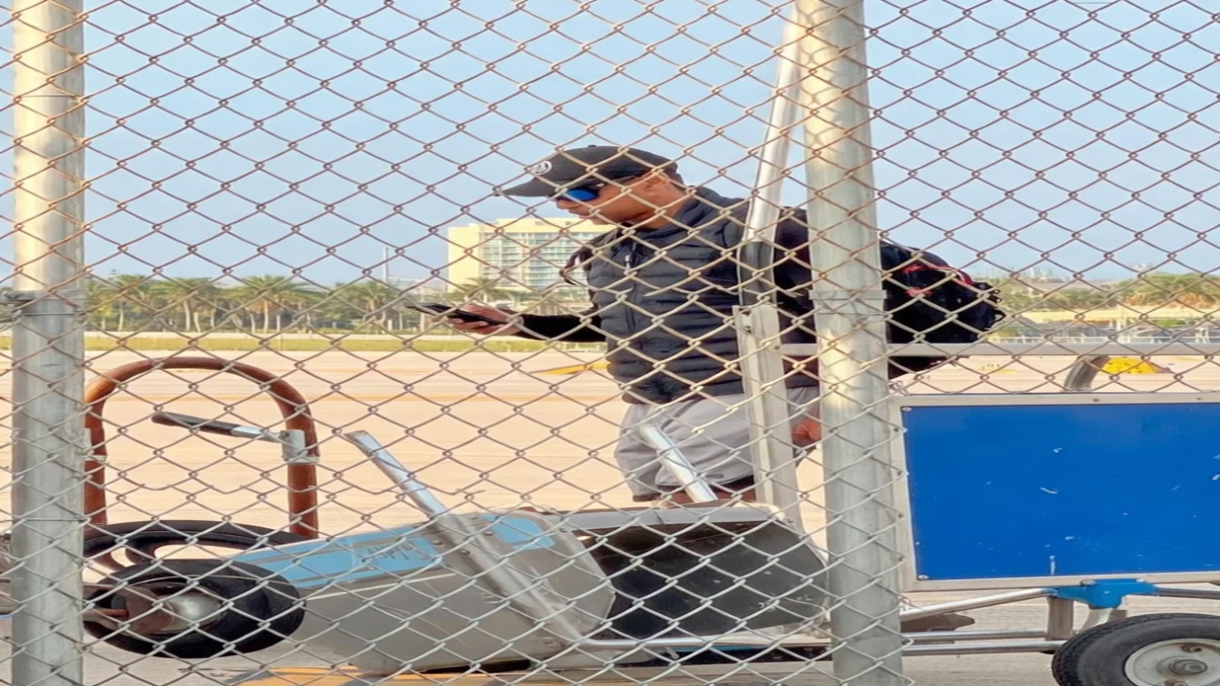 Tiger Woods at an airport, seen through a chain-link fence, wearing sunglasses, a black baseball cap, black jacket, and gray shorts, holding a phone.