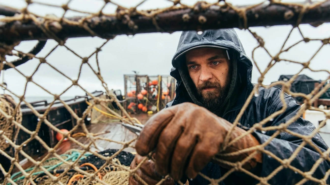 Todd Meadows, a man with a beard and hooded rain jacket, stands on a boat with fishing equipment.