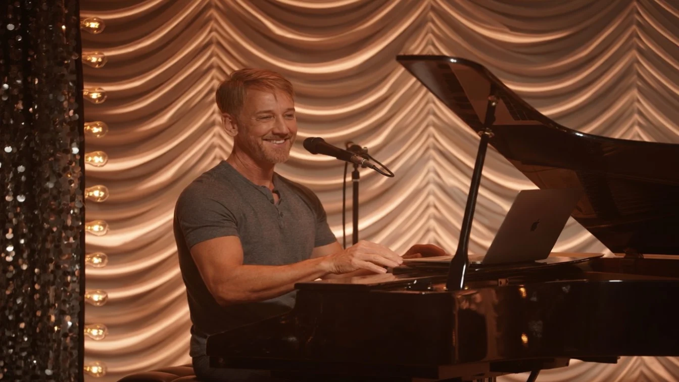 A man with a beard, wearing a grey shirt, smiles as he plays the piano in a spotlight.
