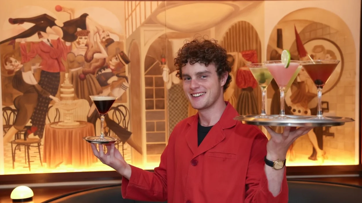 Actor Cameron Scott Roberts in a red jacket holding trays of cocktails.