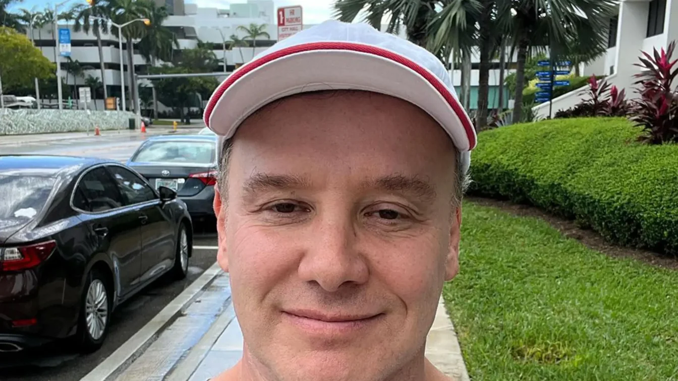 Lenny Hochstein wearing a gray t-shirt with an "I Voted!" sticker, a white hat with red trim, and standing outdoors on a sidewalk next to a street with cars parked.