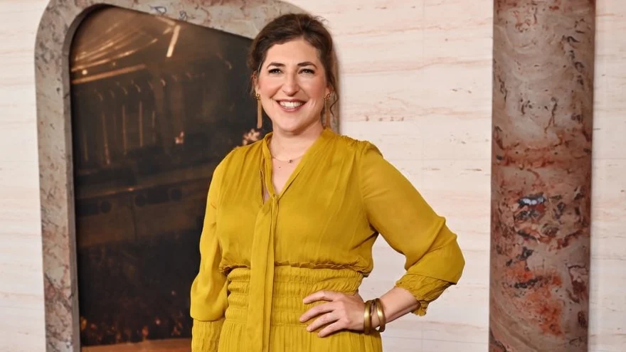 Mayim Bialik in a yellow dress and black boots on a red carpet.
