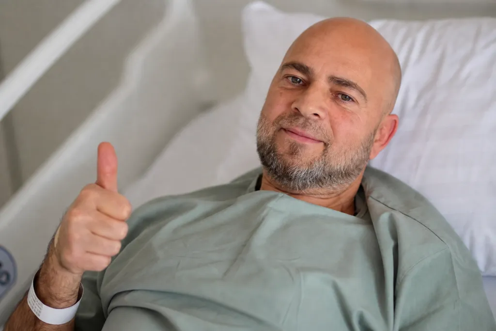 A man giving a thumbs up while lying in a hospital bed wearing a green gown