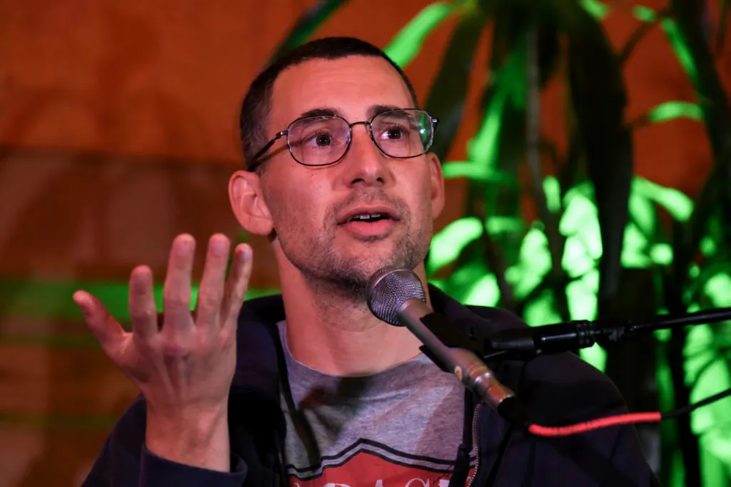 Jack Antonoff speaking into a microphone while sitting and gesturing with his hand in front of plants